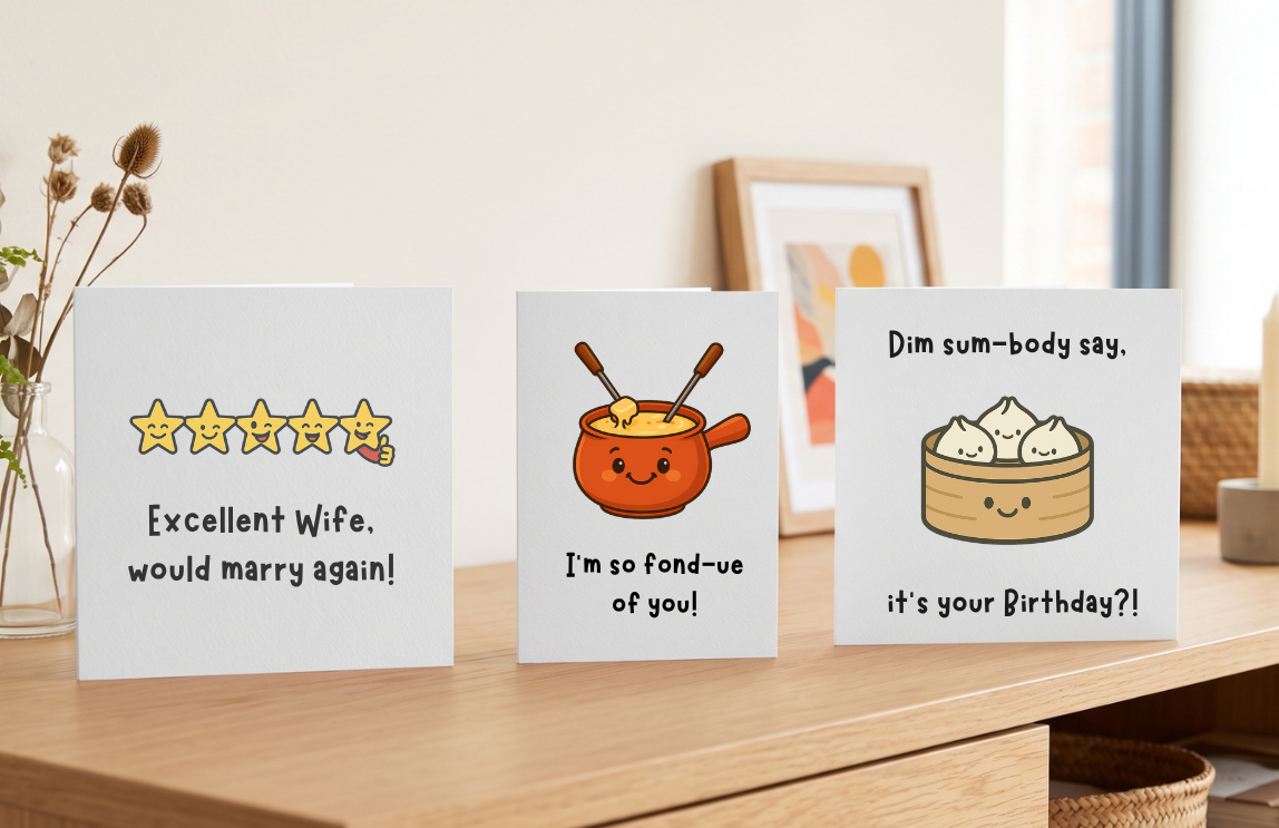 Three funny pun greeting cards standing on a wooden desk beside dried flowers and a framed print